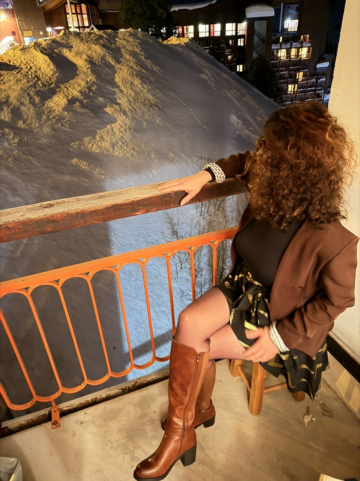 Léna assise le soir sur une chaise, sur le balcon d'un chalet, en blazer et bottes, face au village de ski